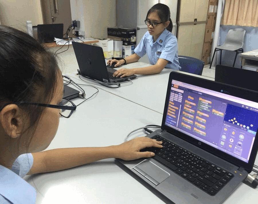 Two students in blue uniforms work on laptops at a white table, one screen showing a colorful coding interface.